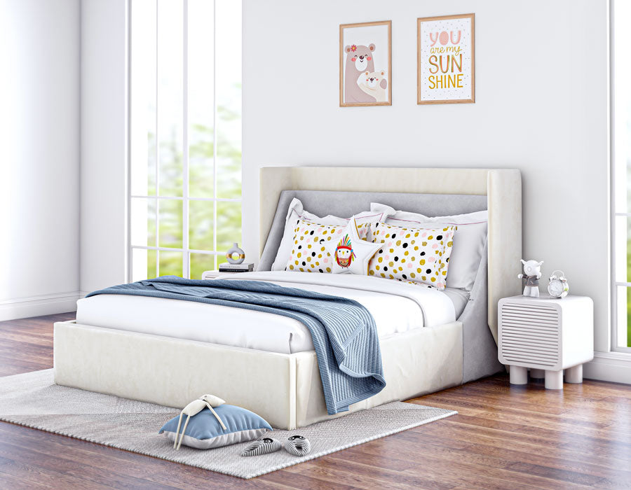 Bright and cozy children's bedroom featuring a cream-colored upholstered bed with a high headboard. The bed is adorned with playful pillows, including a polka dot pattern and a toy. The room has large windows letting in natural light, a white bedside table with decorative items, and wooden flooring. Wall art with positive messages adds a cheerful touch to the inviting space.