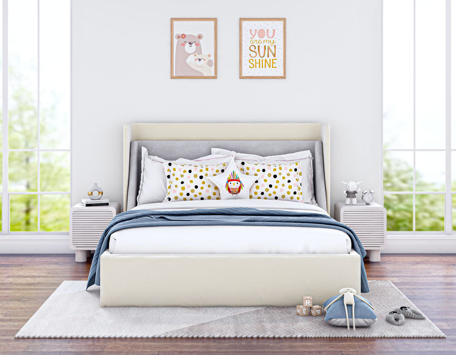 Bright and cozy children's bedroom featuring a cream-colored upholstered bed with a high headboard. The bed is adorned with playful pillows, including a polka dot pattern and a toy. The room has large windows letting in natural light, two white bedside tables with decorative items, and wooden flooring. Wall art with positive messages adds a cheerful touch to the inviting space.